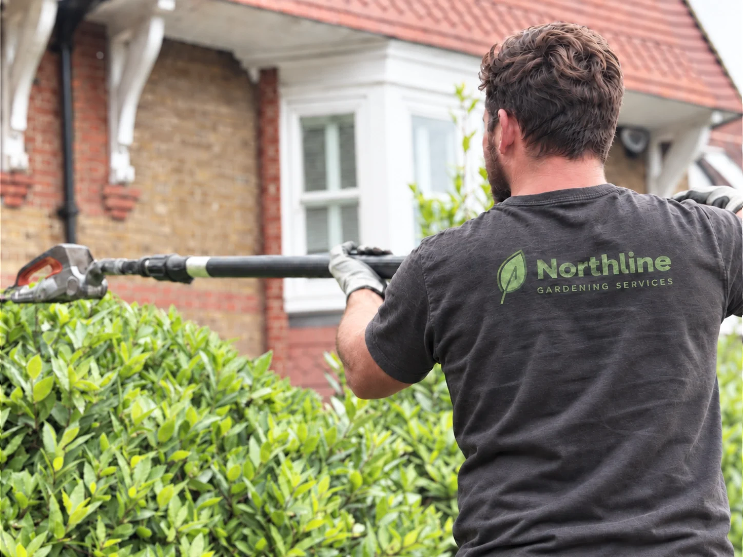 Northline Gardening Services workwear: the leaf and horizontal wordmark printed in green on the back of a grey t-shirt worn by a gardener trimming a hedge